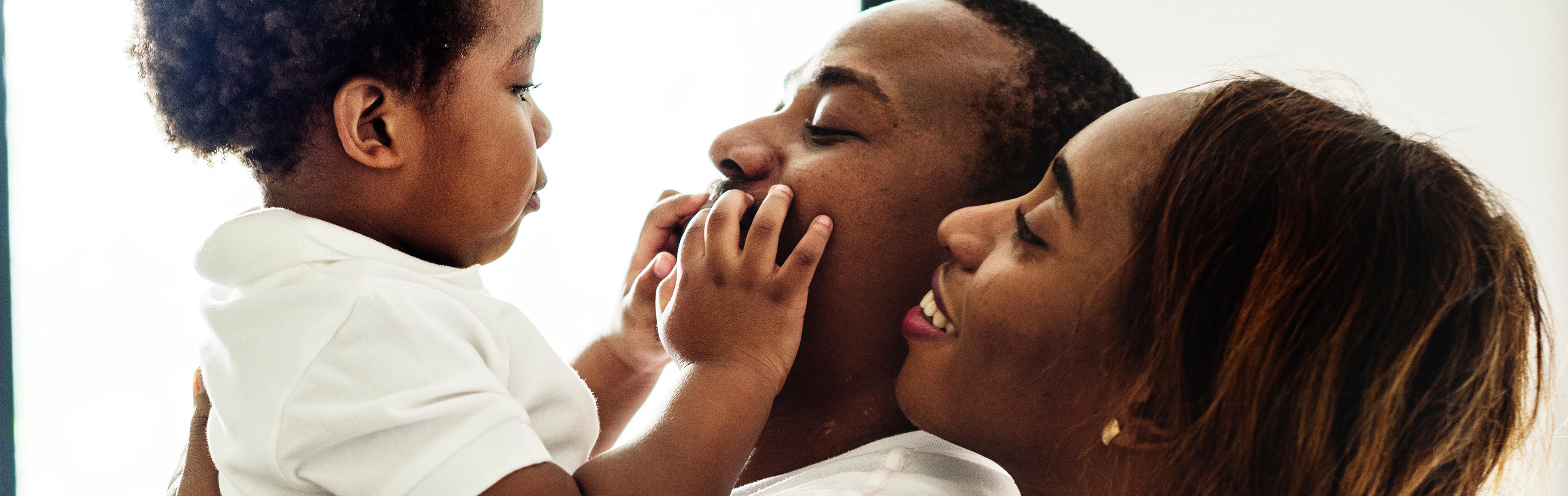 Father and mother holding the baby up while the baby is playing with the father's mouth.
